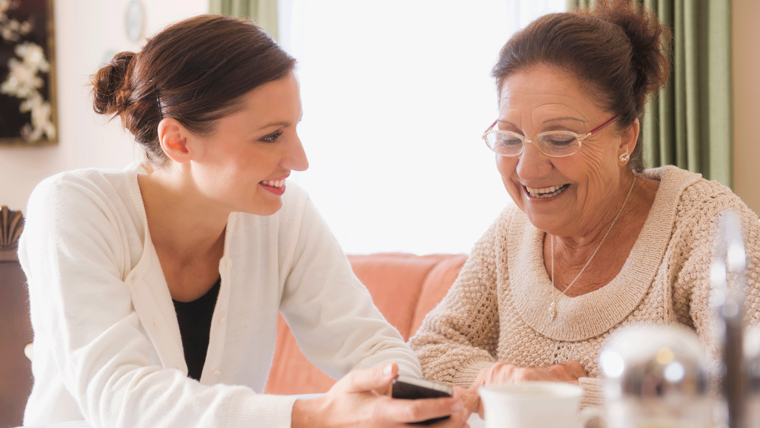 An older woman and a younger woman smile as they look at a phone together, sharing a moment.