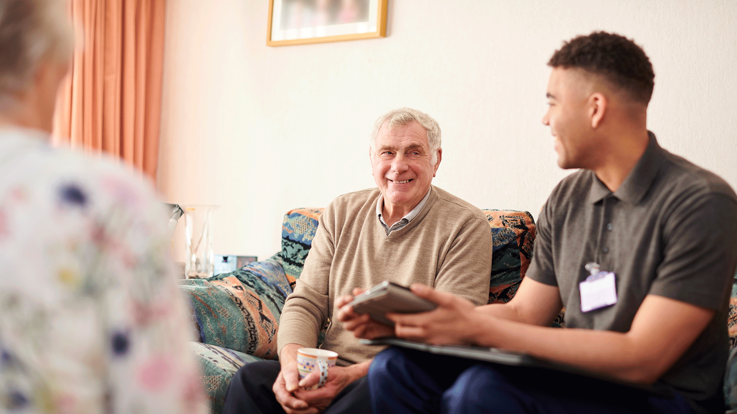 A caregiver and an elderly person sit on a sofa in an aged care facility, discussing something while drinking tea together.