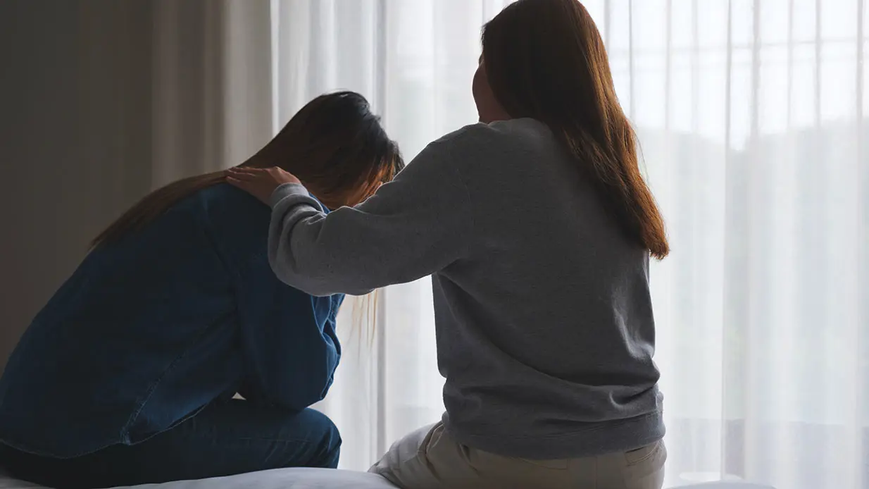 Two women sitting on a bed, one comforting the other