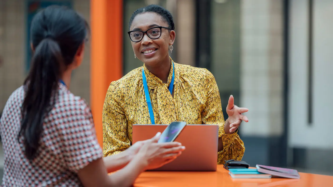 A teacher talking with her colleague in a school hall with her laptop open on a nearby table.