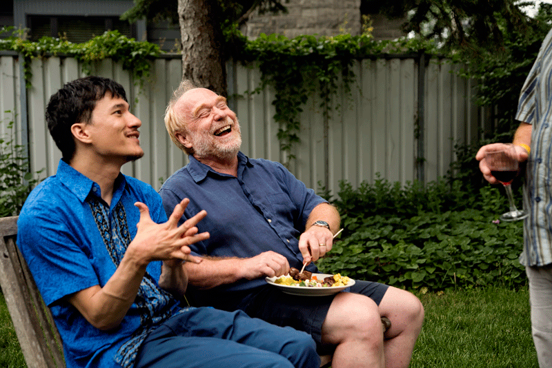 Two men smiling and laughing outdoors while sitting on a bench, enjoying a casual conversation.