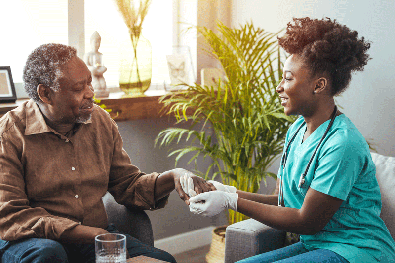 A nurse in scrubs smiles while holding the hand of an elderly man in a warmly lit room with plants.