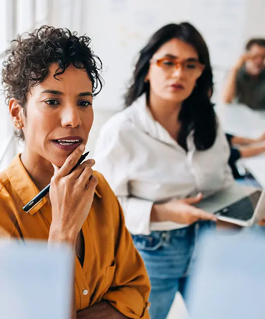 A woman holding a pen and another woman holding a laptop contemplate a problem. 