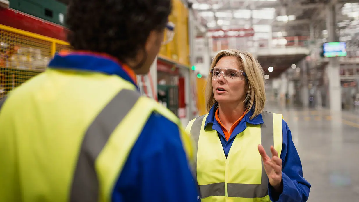 Two people in safety gear talking in a warehouse.