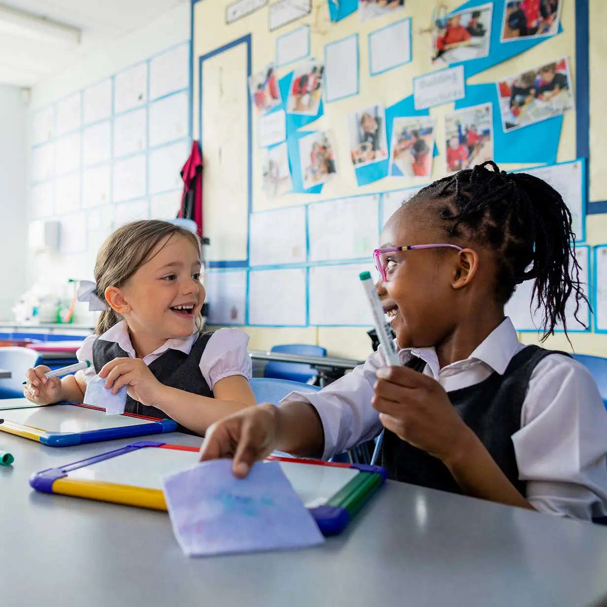 Two elementary school students at a table writing on whiteboard tablets.