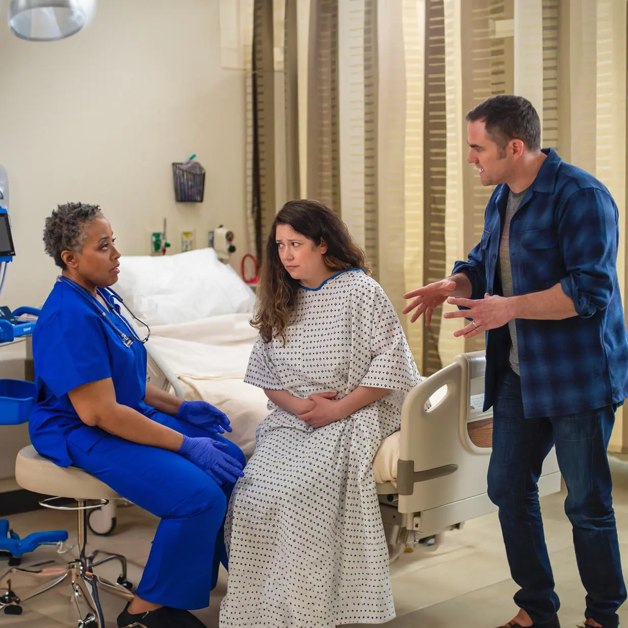 An angry man talking to a medical professional while a female patient sits on the bed looking on.